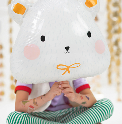 Balão em forma de rosto de urso branco com detalhes amarelos e cor-de-rosa, segurado por criança sentada com roupa colorida