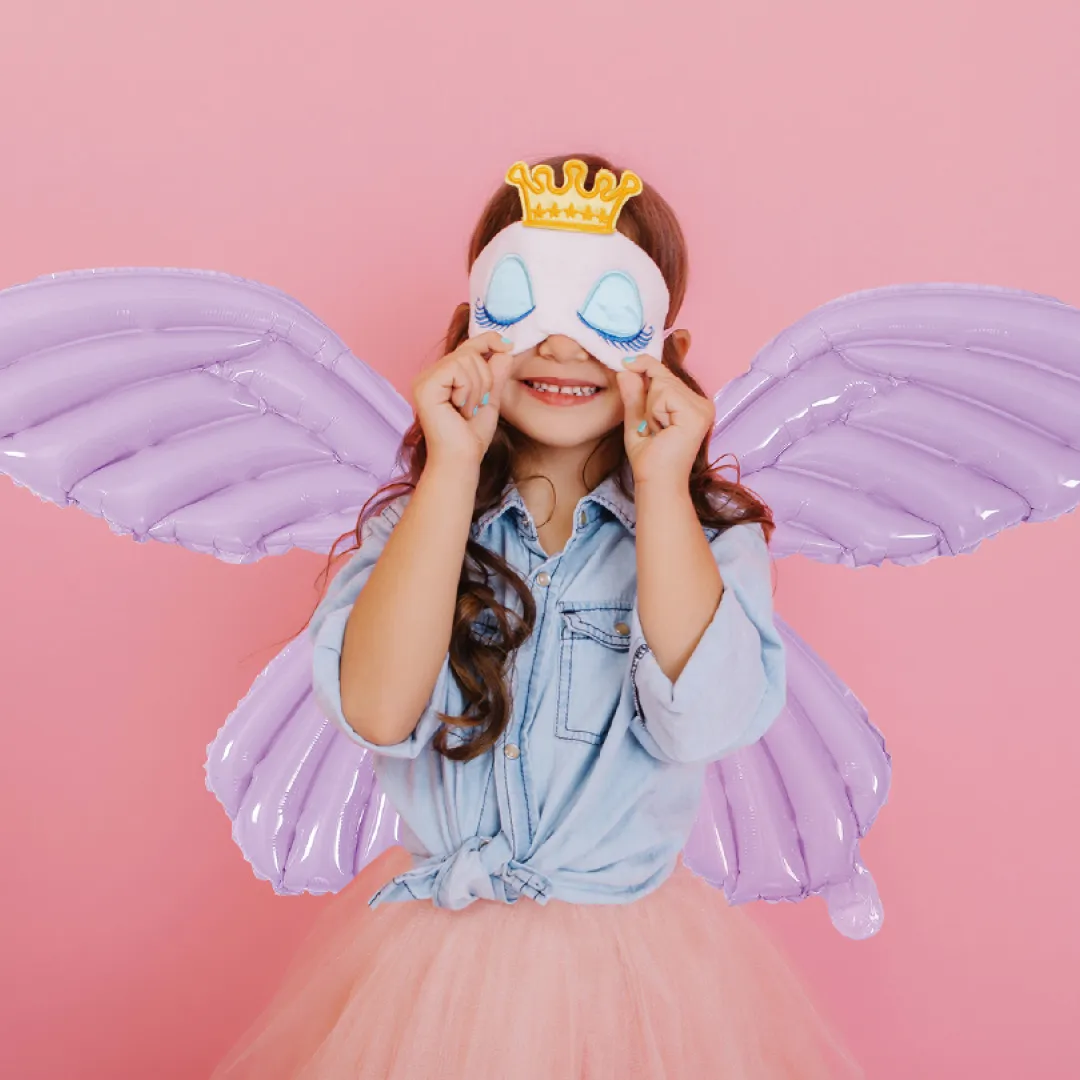 Menina com asas infláveis lilás, máscara branca e coroa dourada num fundo rosa