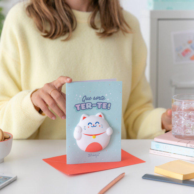 Cartão azul com gato da sorte tridimensional e texto em português sobre mesa branca