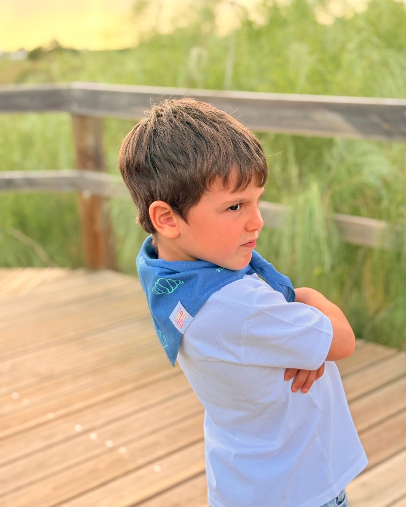 Criança de perfil com camisola branca e bandana azul com padrão de conchas em passadiço de madeira