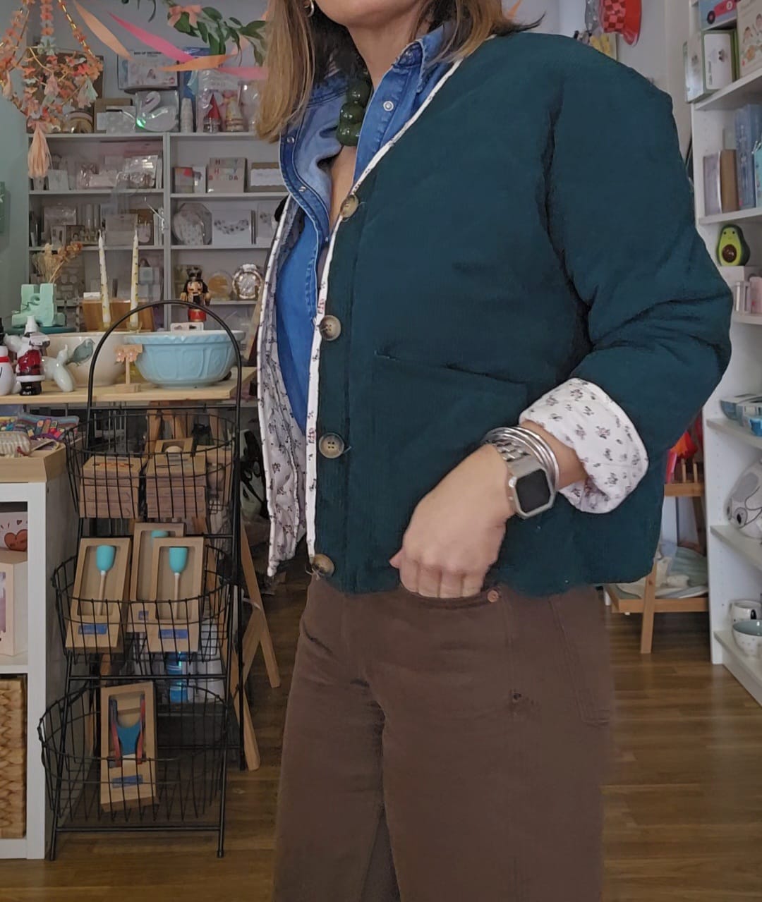 Pessoa vestindo casaco verde-escuro com botões e camisa azul, com relógio e braceletes prateados, em loja com prateleiras e diversos objetos.