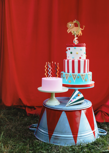 Três bolos coloridos decorados sobre suporte metálico com fundo de cortina vermelha