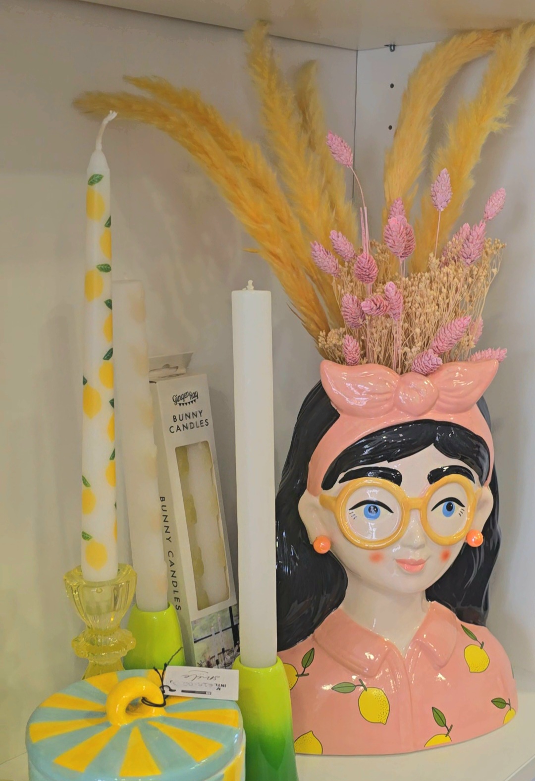 Vaso decorativo cerâmico com rosto feminino e flores secas, velas e embalagens ao lado.