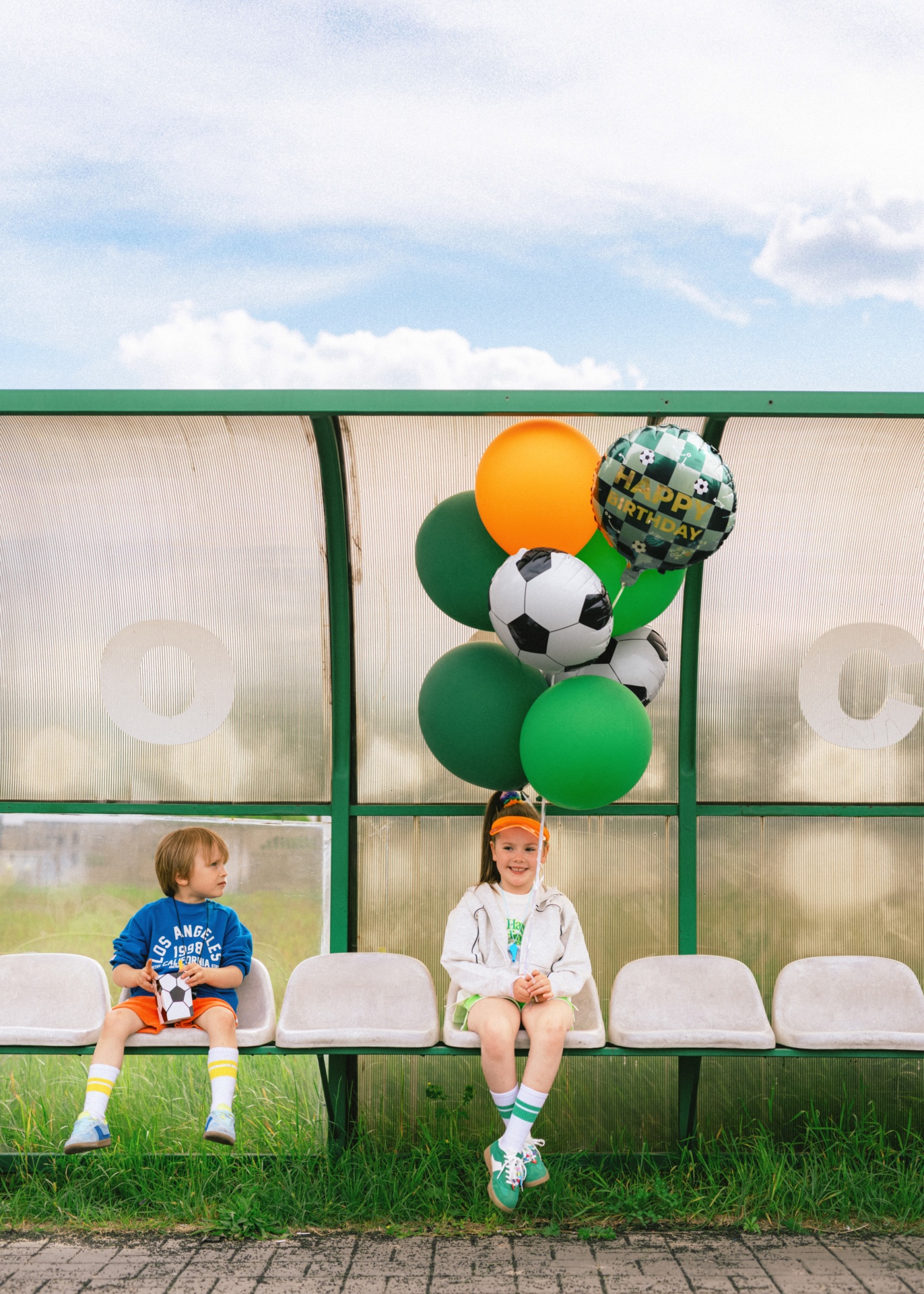 Duas crianças sentadas num banco ao ar livre, uma com balões de futebol e um balão de feliz aniversário