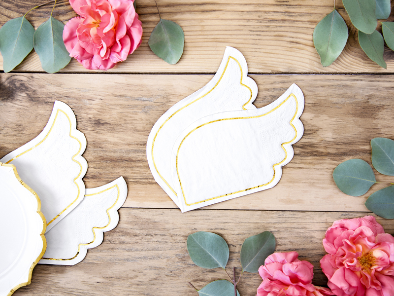 Guardanapos em forma de asas brancas com bordas douradas sobre mesa de madeira com flores e folhas verdes