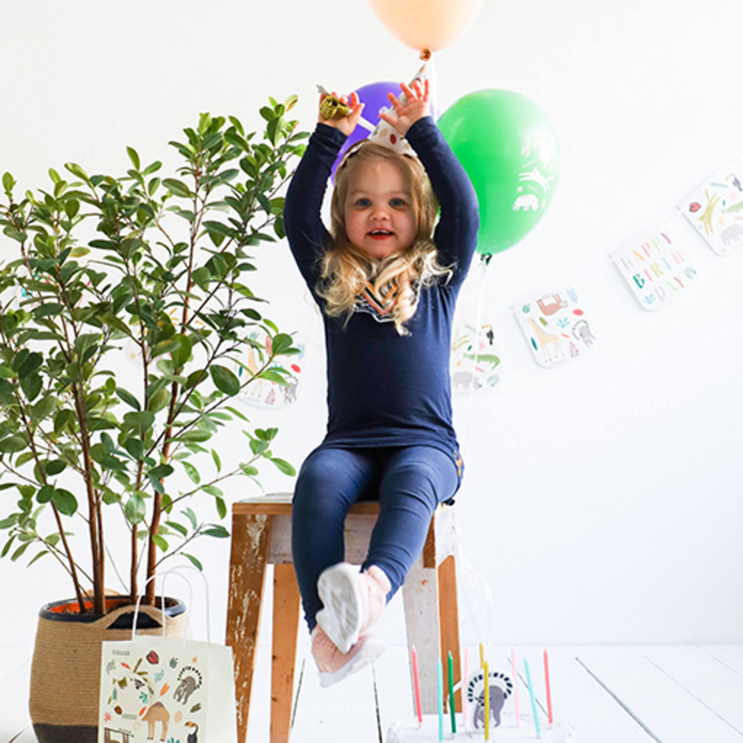 Menina com pijama azul segurando balões coloridos sentada numa cadeira de madeira com decoração de aniversário ao fundo