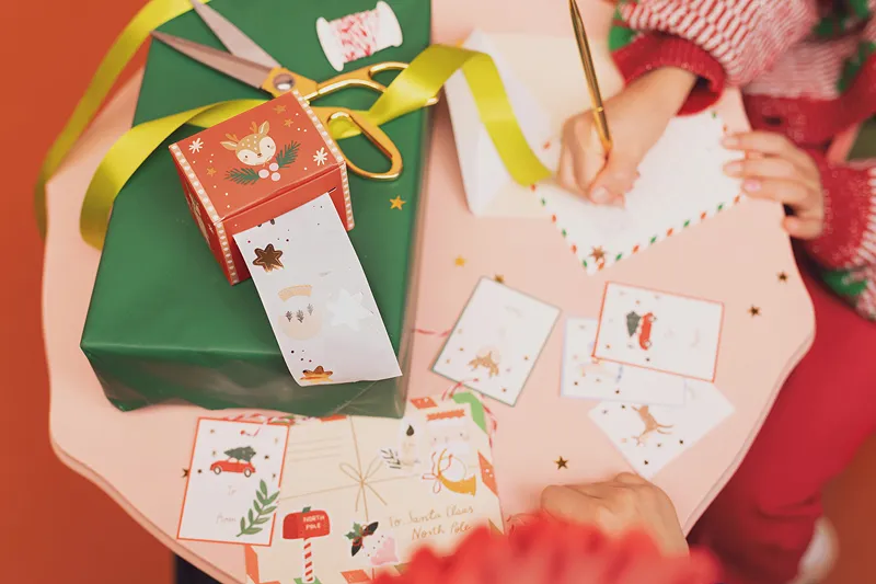 Mãos a escrever num cartão de Natal com fita decorativa e papel de presente verde sobre uma mesa rosa.