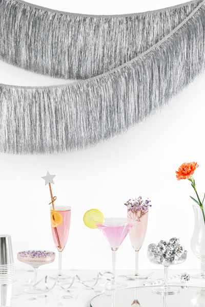 Quatro taças com bebidas rosa e decoração, franjas prateadas na parede, vaso branco com flor laranja