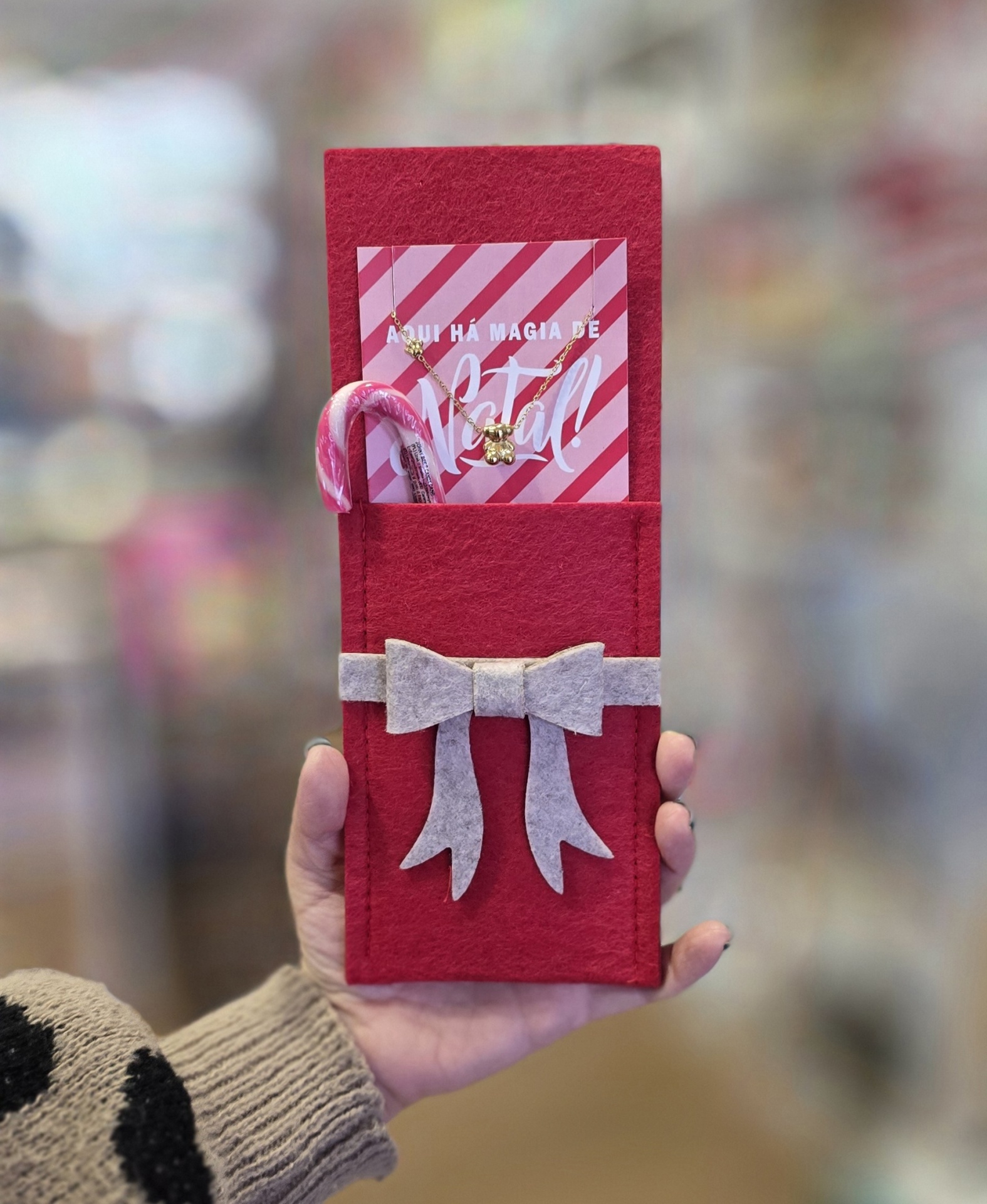 Embalagem vermelha de feltro com laço cinzento, cartão rosa, colar dourado e canequinho rosa e branco.