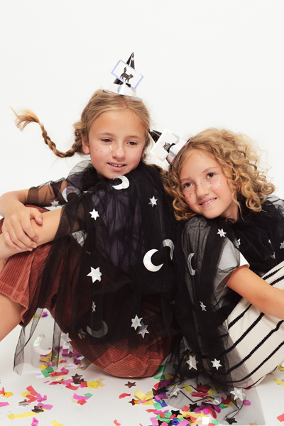 Duas meninas de capa preta de tule com estrelas e luas, chapéus de festa brancos, sentadas em chão com confetes coloridos