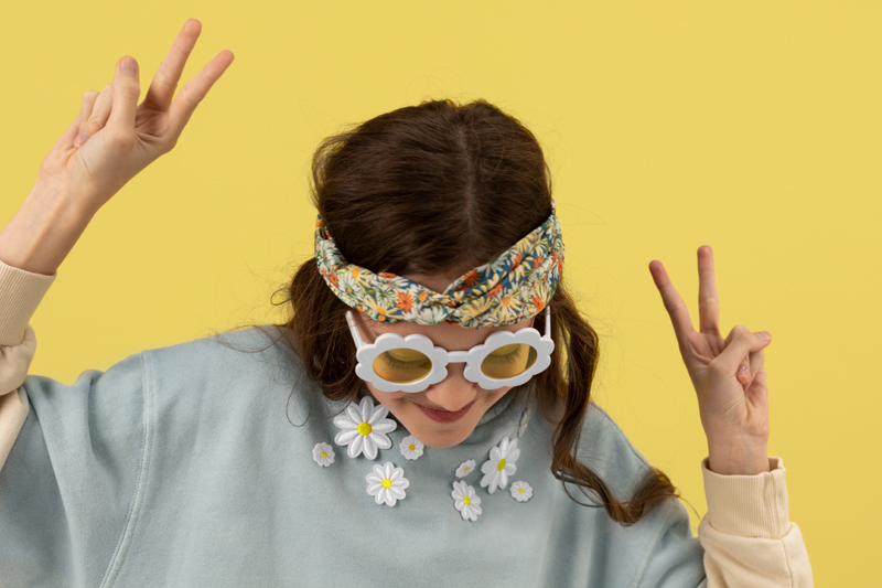 Pessoa com óculos de sol flor branca, faixa de cabeça floral e sweat azul claro com flores, fundo amarelo.