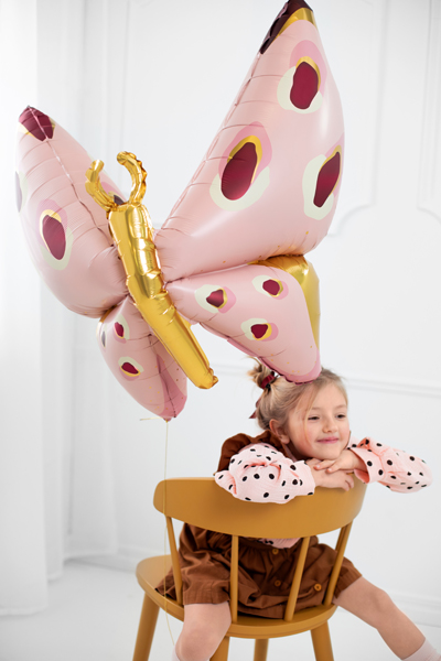 Balão metálico em forma de borboleta cor-de-rosa com manchas coloridas e corpo dourado amarrado a uma cadeira de madeira com menina sentada