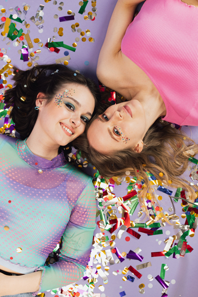 Duas mulheres sorridentes deitadas, vestindo roupas coloridas com confetes ao redor.