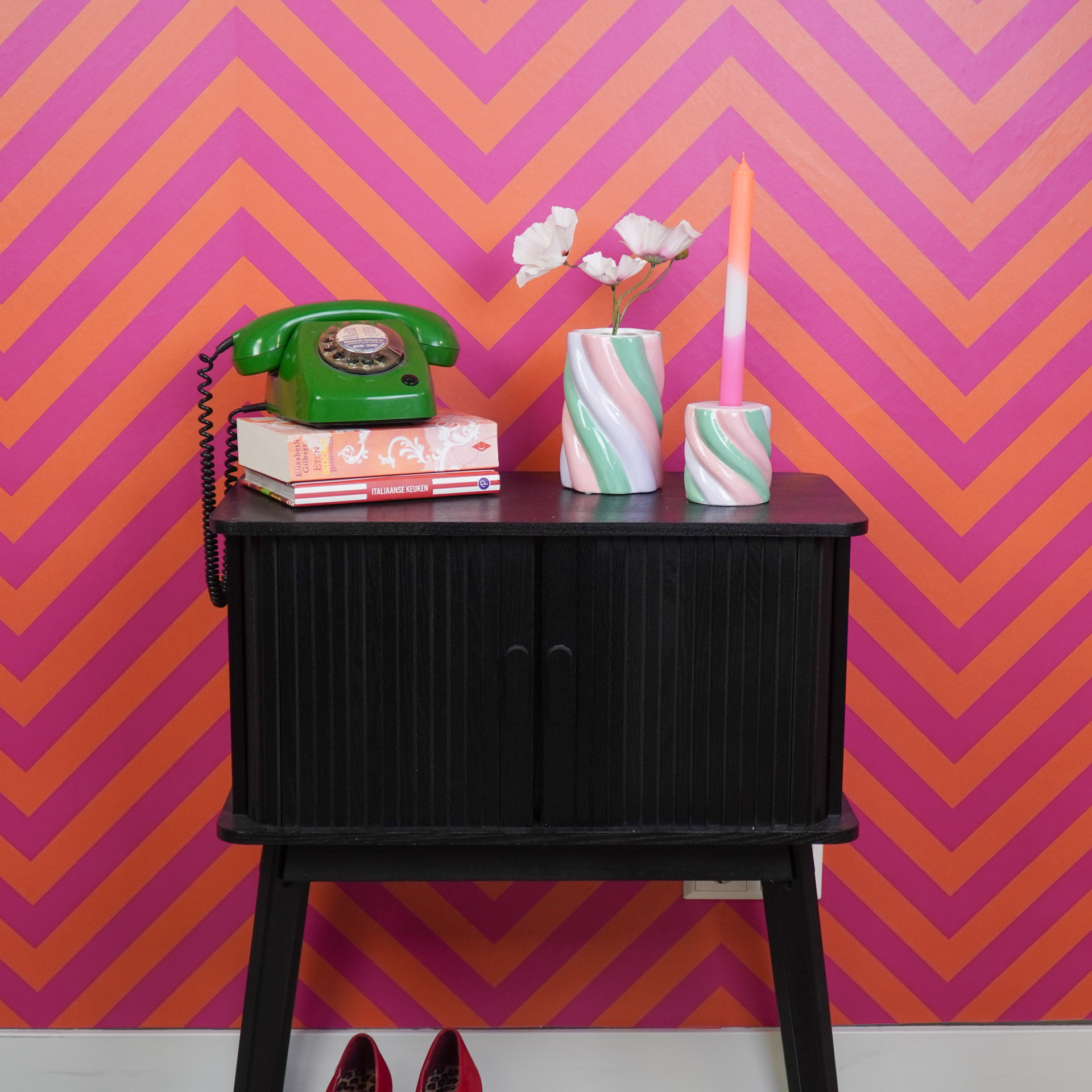 Móvel preto com telefone verde, livros, vaso com flores e castiçal com vela em fundo de parede laranja e rosa
