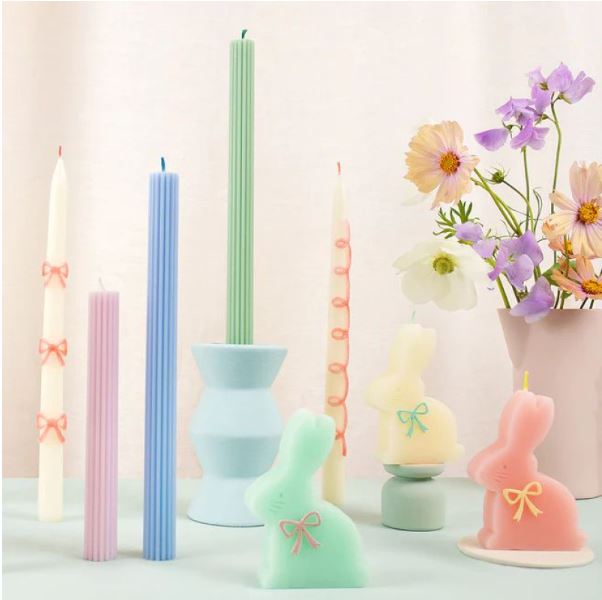 Velas coloridas variadas, incluindo velas altas e velas em forma de coelho, decoradas com laços, com flores em vaso ao fundo.