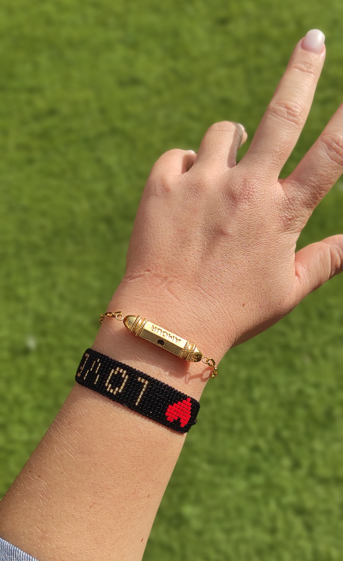 Pulso com duas pulseiras, uma preta com letras douradas e coração vermelho, outra dourada com placa AMOUR, fundo relva verde