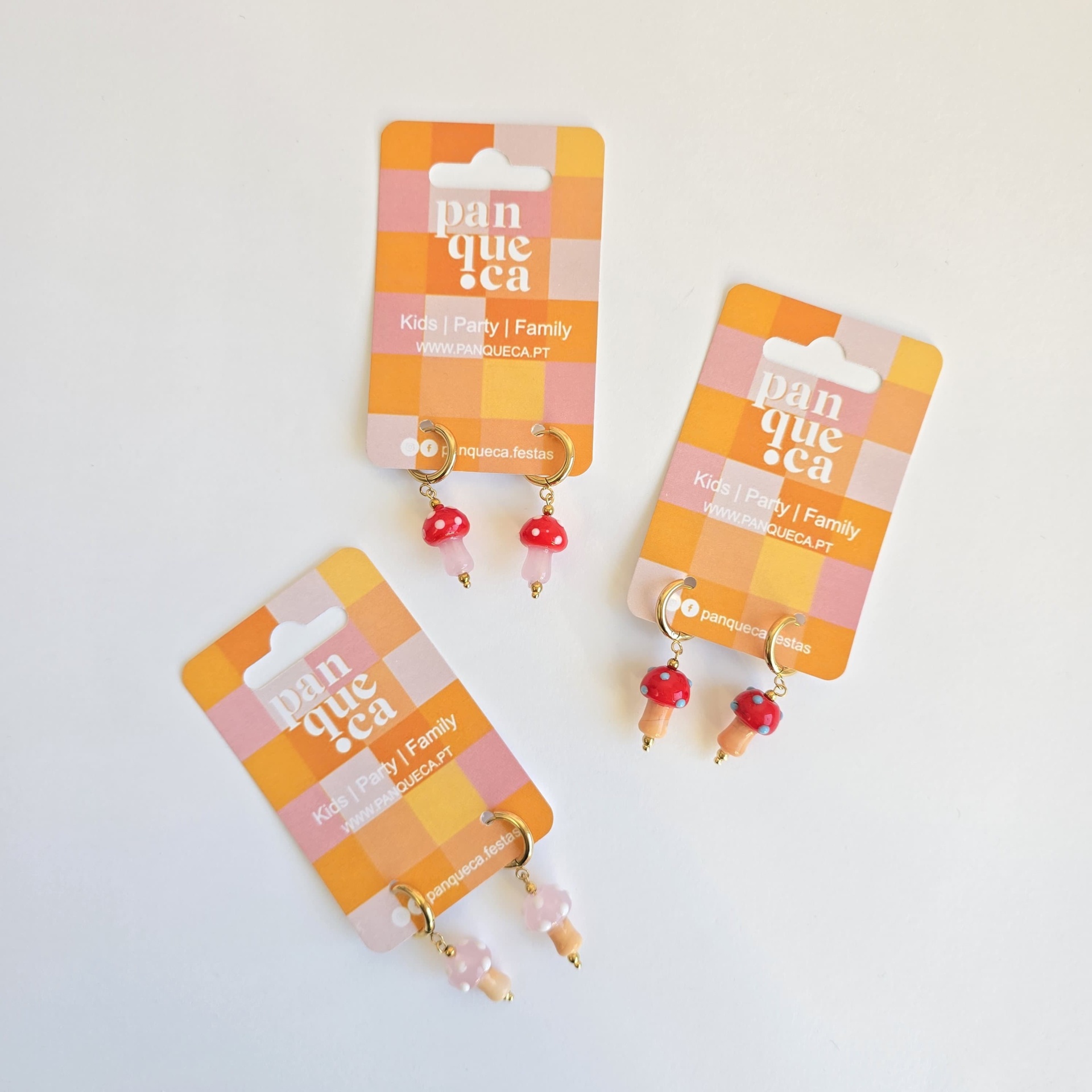 Três pares de brincos coloridos em cartões com padrão xadrez laranja e rosa da marca Panqueca.