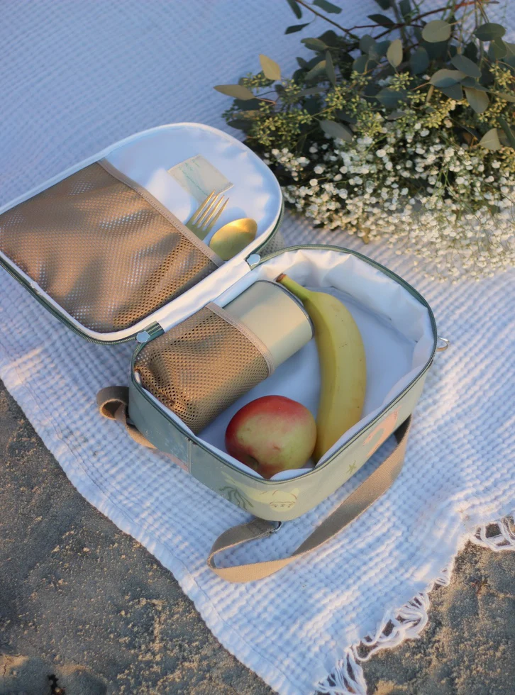 Mala térmica cinza claro aberta com frutas e talheres, sobre manta branca e flores.