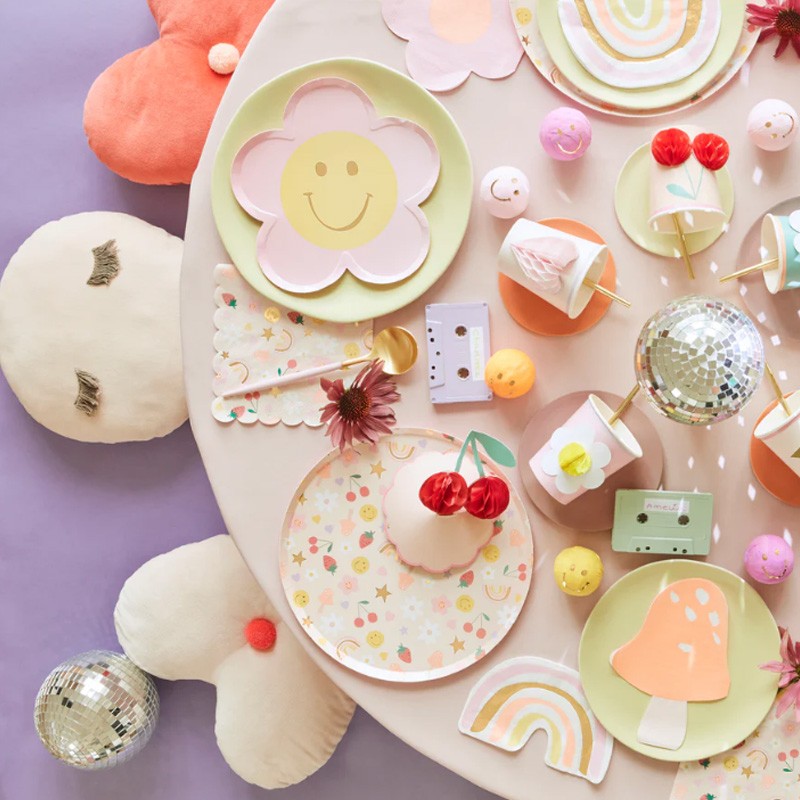 Mesa decorada com pratos coloridos, copos com flores e cerejas, esferas espelhadas e almofadas de flores