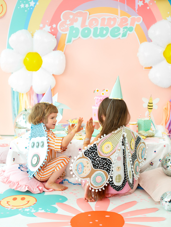 Crianças com asas e chapéus em festa com decoração floral e arco-íris 'Flower power'
