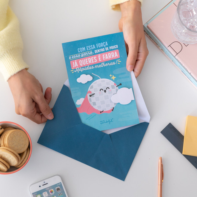 Cartão azul com ilustração de bola sorridente e texto em português, mãos a colocar dentro de envelope azul numa mesa com objetos