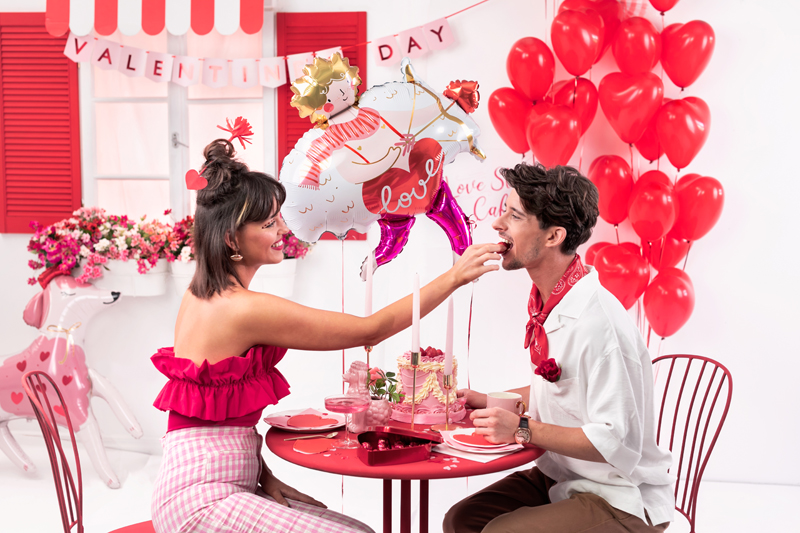 Casal a partilhar comida numa mesa com decoração do Dia dos Namorados com balões vermelhos e faixa com 'VALENTINE'S DAY'