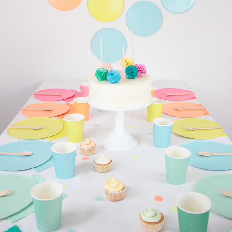 Mesa de festa com bolo branco, cupcakes coloridos, pratos, copos e colheres em tons pastel e decoração de círculos coloridos na parede