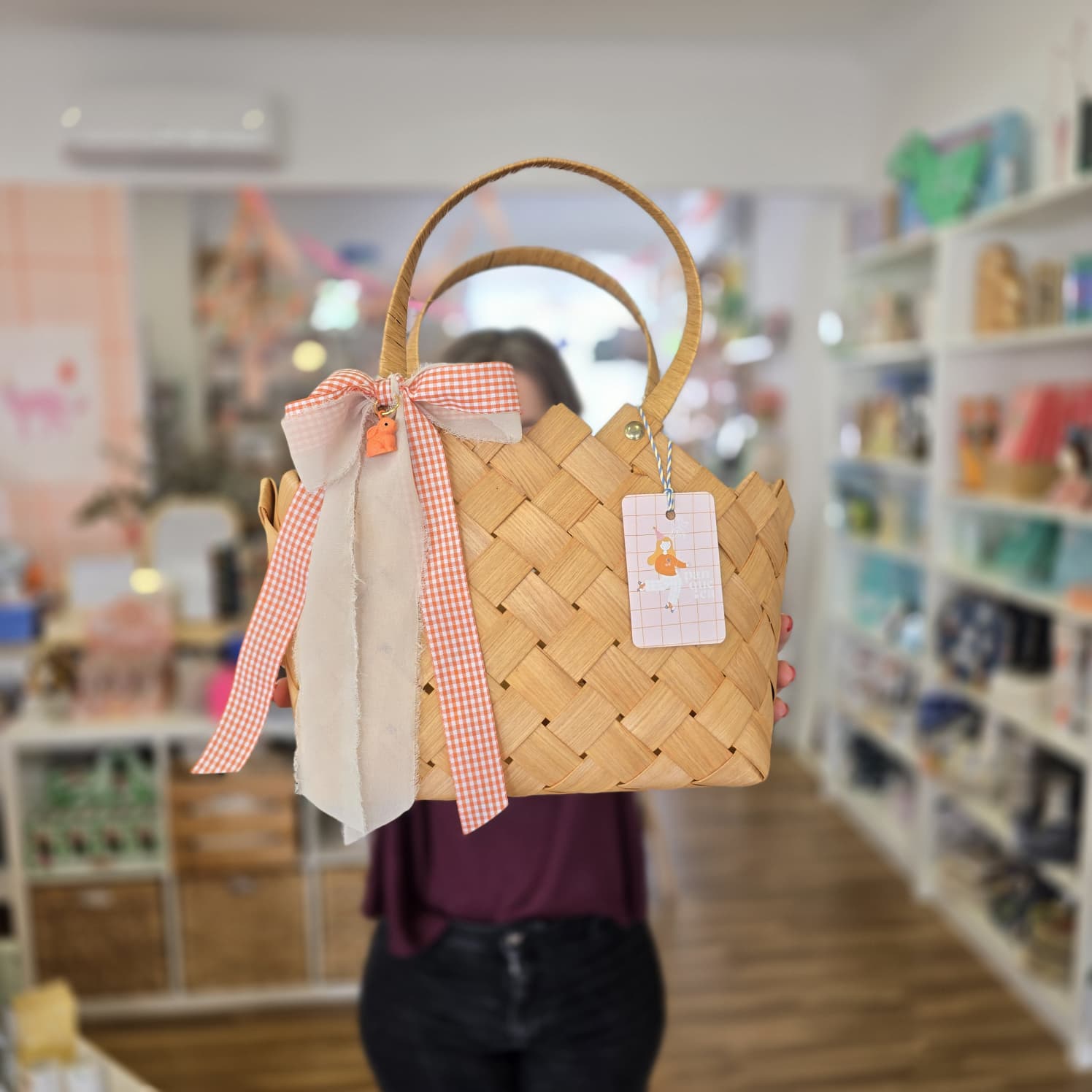 Bolsa de mão em palha com laço xadrez laranja e bege e etiqueta pendurada, em loja desfocada.