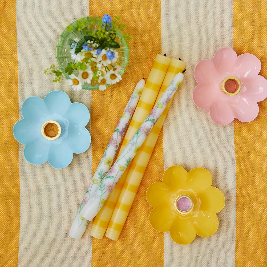 Velas decorativas finas florais e às riscas com suportes de velas em forma de flor e vaso com flores sobre tecido às riscas amarelo e bege.