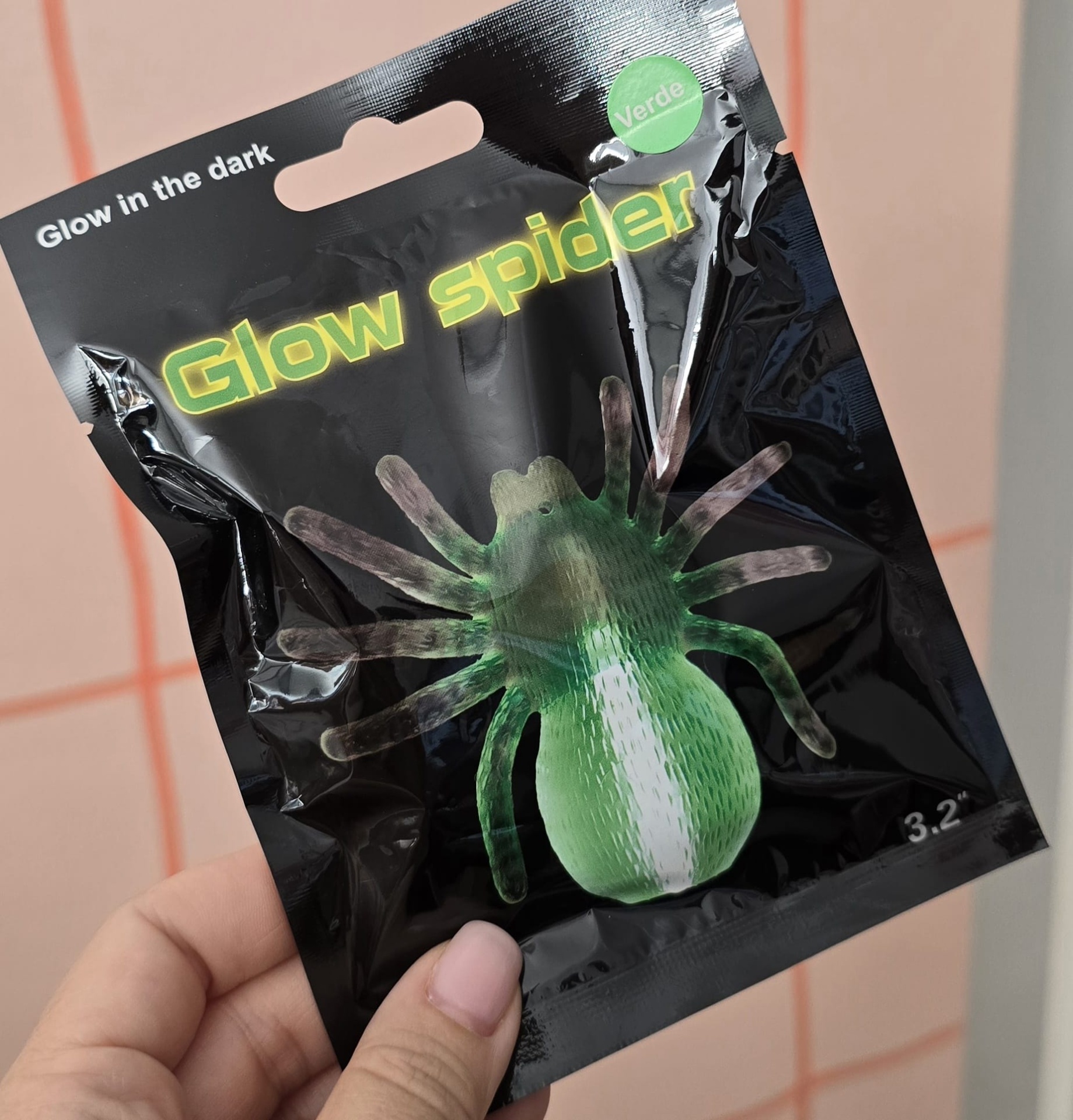 Embalagem preta brilhante com brinquedo verde de aranha, textos em inglês e português, mão segurando.