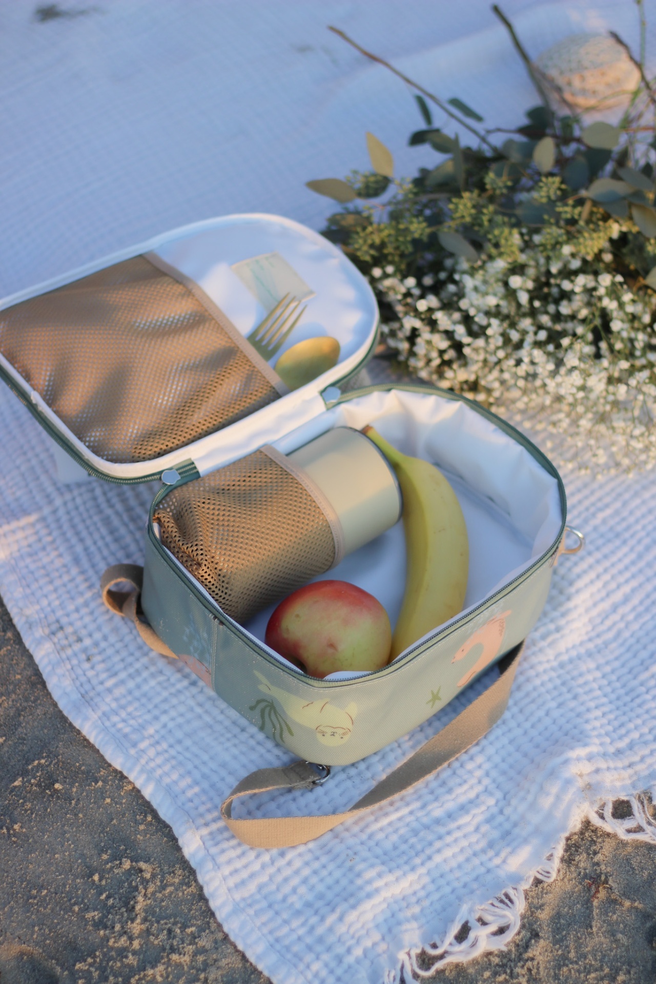 Estojo térmico verde claro com frutas e chávena, aberto sobre manta branca na areia