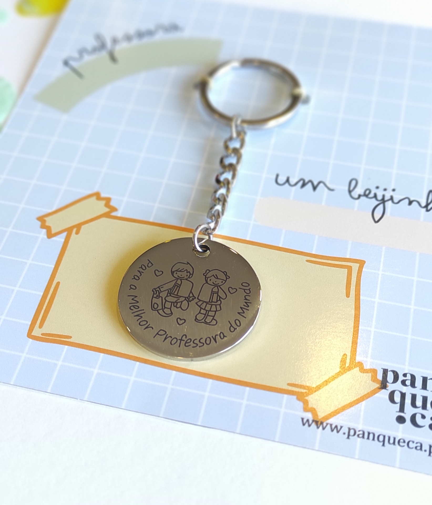 Chaveiro metálico redondo prateado com gravação e texto em português pendurado em correntinha sobre cartão quadriculado.