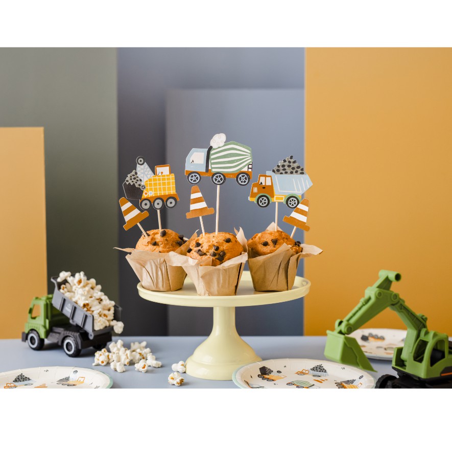 Muffins decorados com toppers de veículos de construção sobre base creme com brinquedos e pratos temáticos ao redor