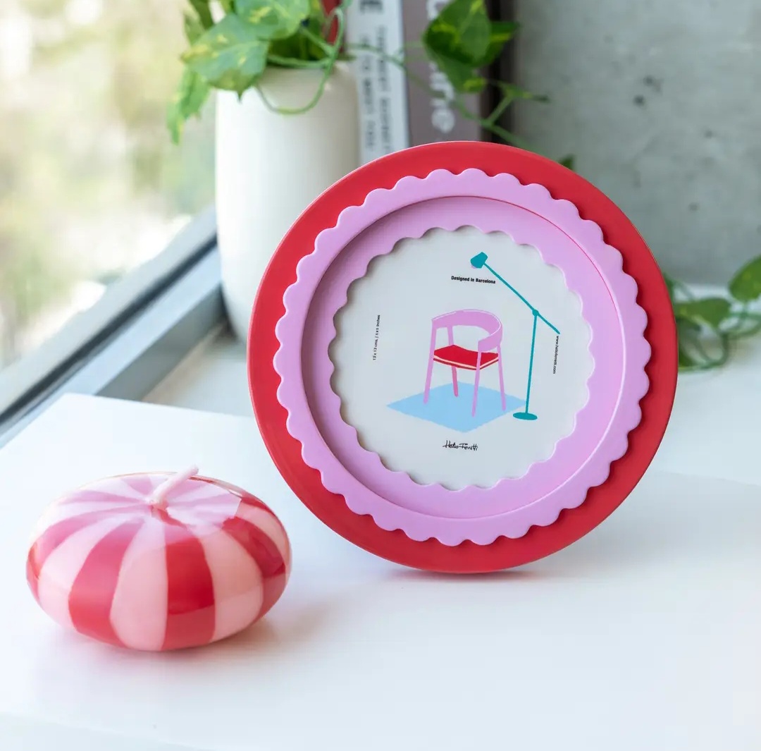 Suporte de cera vermelho e rosa com design de cadeira e candeeiro ao lado de uma vela redonda às riscas vermelhas e brancas, junto a janela.