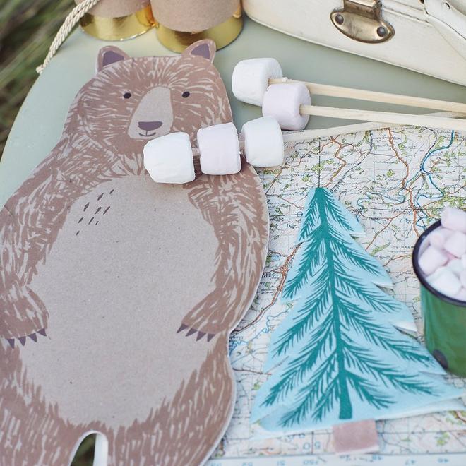 Almofada com estampa de urso, marshmallows em espetos, mapa com pinheiro azul e mala branca