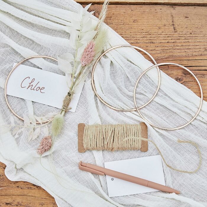 Dois anéis dourados, raminho de flores secas, rolo de fio corda, caneta rosa e etiquetas sobre tecido branco e mesa de madeira