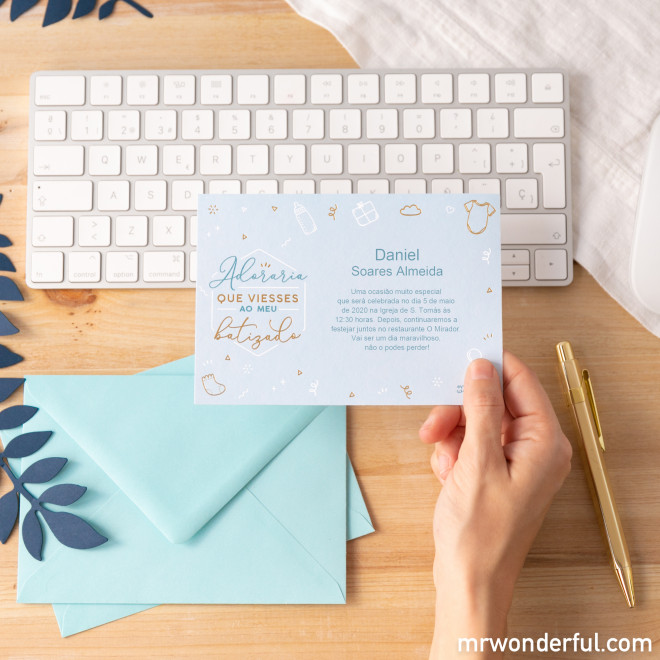 Convite azul claro para batizado com texto e decoração dourada, junto a teclado branco, envelopes azuis e caneta dourada