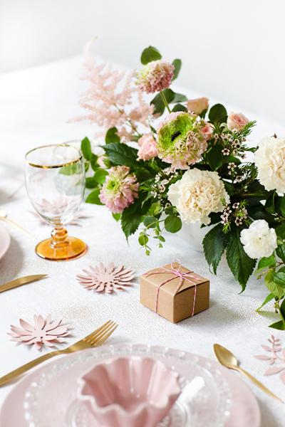 Mesa posta com pratos rosa, talheres dourados, copo âmbar, arranjo floral e caixa de presente
