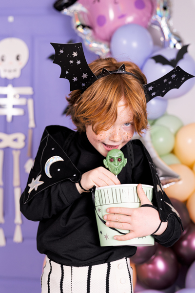 Criança vestida com blusão preto decorado e tiara de morcego segura balde com objeto verde