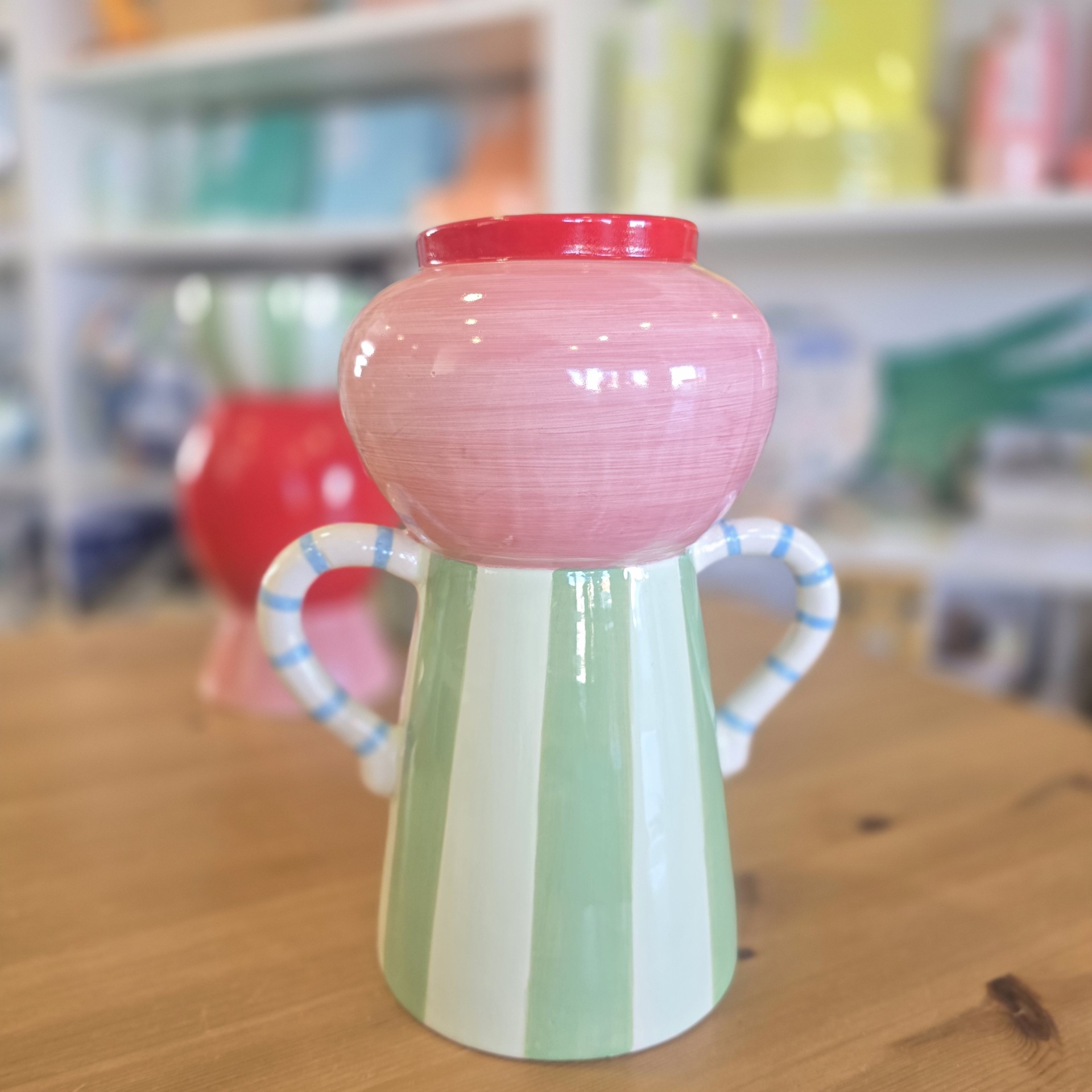 Jarro de cerâmica verde, branco e rosa com pegas listradas azuis numa mesa de madeira