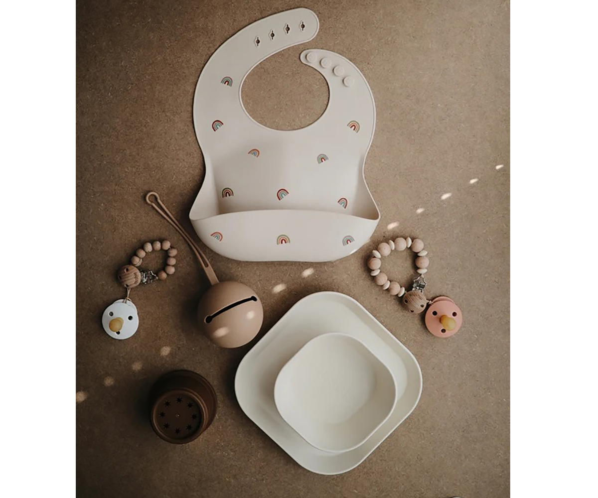 Babete em silicone creme, pratos brancos, mordedores de madeira e chocalho sobre superfície castanha