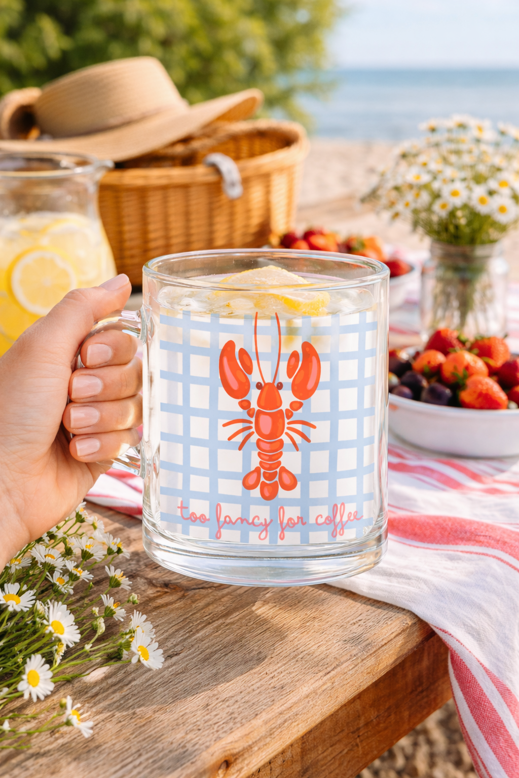 Caneca de vidro com lagosta vermelha e texto too fancy for coffee numa mesa de madeira com flores e frutas, praia ao fundo.