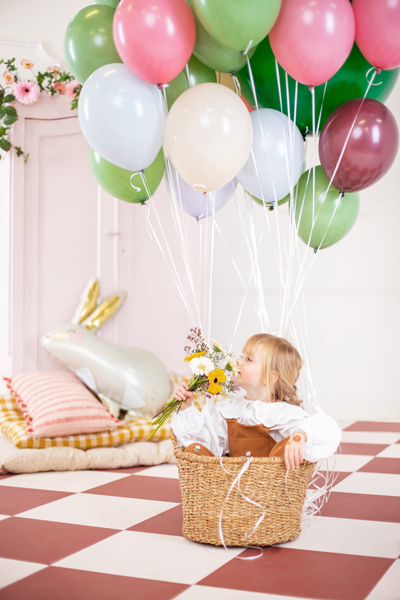 Criança numa cesta de verga com balões coloridos segurando flores em ambiente decorado.