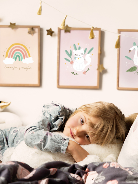 Criança de pijama deitada numa cama branca, com quadros decorativos de arco-íris, gato e cisne ao fundo
