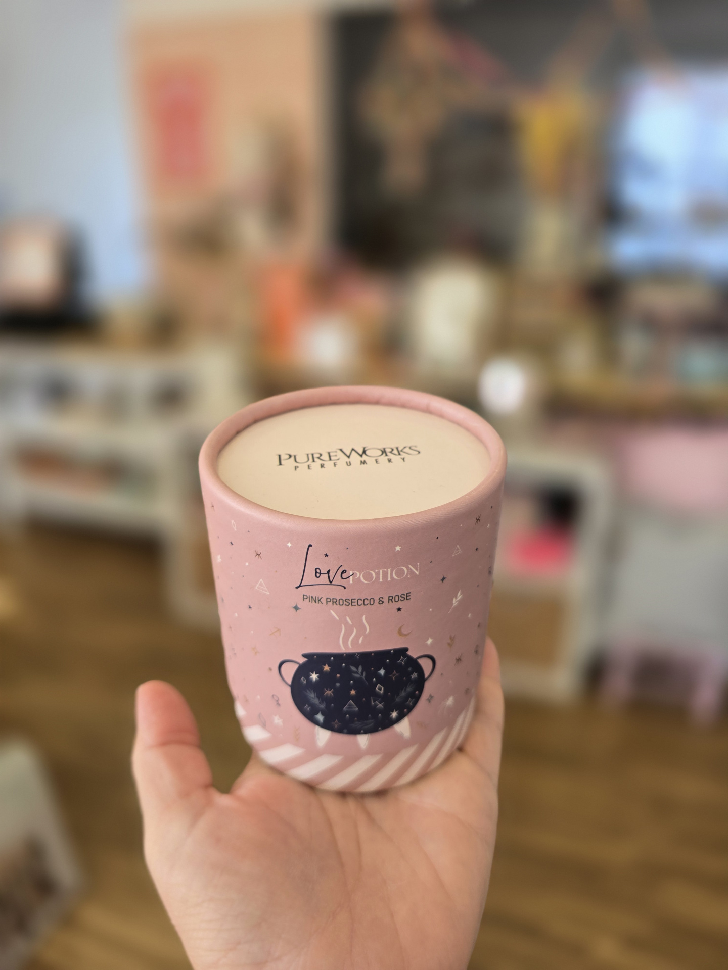 Recipiente de vela ou perfume rosa claro com tampa creme e ilustração de pote preto, segurado numa mão.