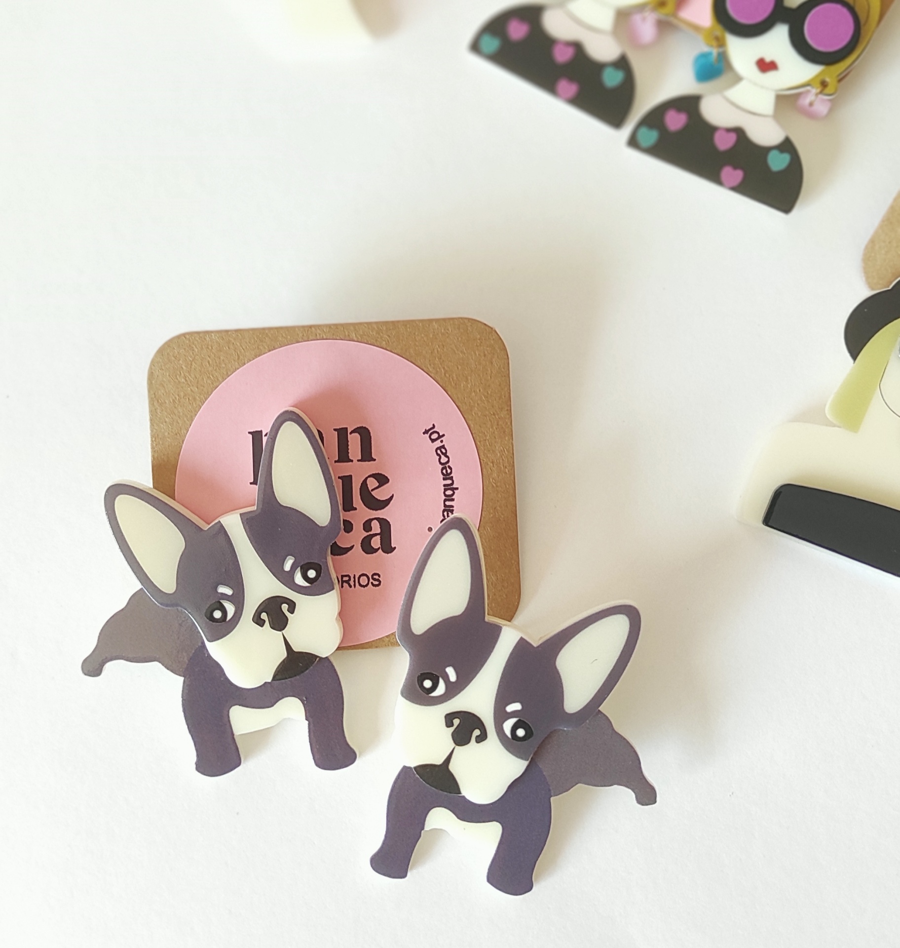 Brincos de bulldog francês em plástico preto, branco e cinza sobre cartão castanho com etiqueta rosa