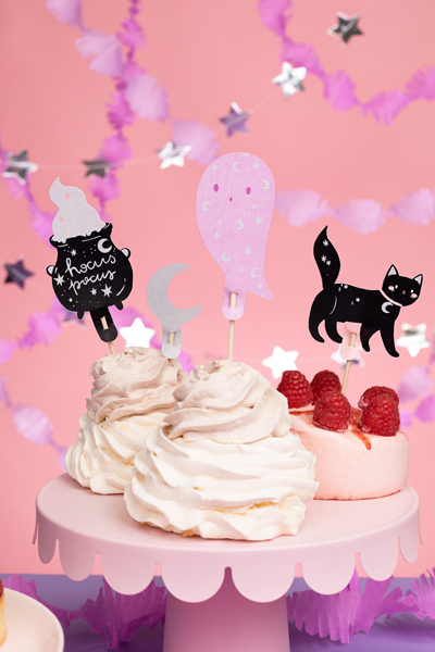 Bolo decorado com chantilly branco e rosa, framboesas frescas e toppers de papel com desenhos de caldeirão, lua, figura rosa e gato preto sobre suporte rosa