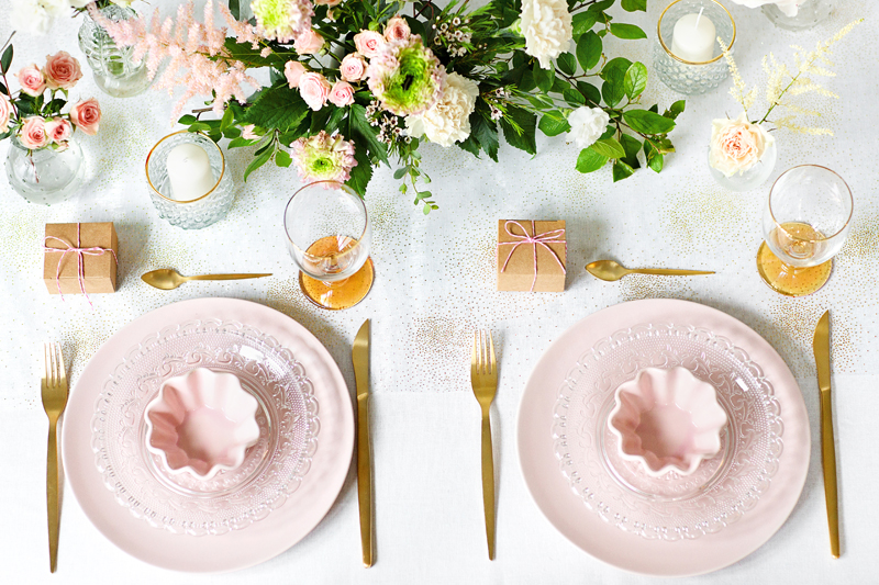 Mesa posta decorada com pratos rosas, talheres dourados, taças e flores