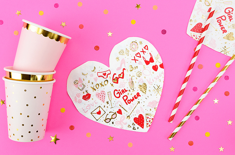 Copos de papel brancos com detalhes dourados e decoração 'Girl Power' sobre fundo rosa com confetes