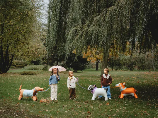 Crianças a passear cães insufláveis coloridos num parque com árvores e relva